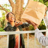 young Black woman hands brown paper bag over the fence to elderly female neighbor